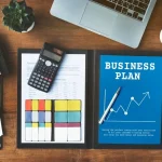 Dokumen Business Plan di atas meja kerja dengan kalkulator, laptop, dan grafik keuangan