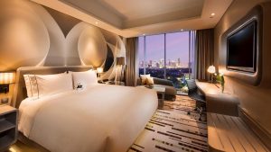 Kamar hotel bintang 5 mewah dengan desain modern, tempat tidur king size, pemandangan kota Jakarta dari jendela besar, serta fasilitas lengkap seperti meja kerja, TV layar datar, dan sofa elegan