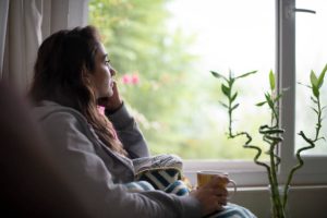 Seorang wanita duduk di dekat jendela, merenung sambil memegang secangkir teh, mencerminkan perenungan tentang cinta dan kenyamanan